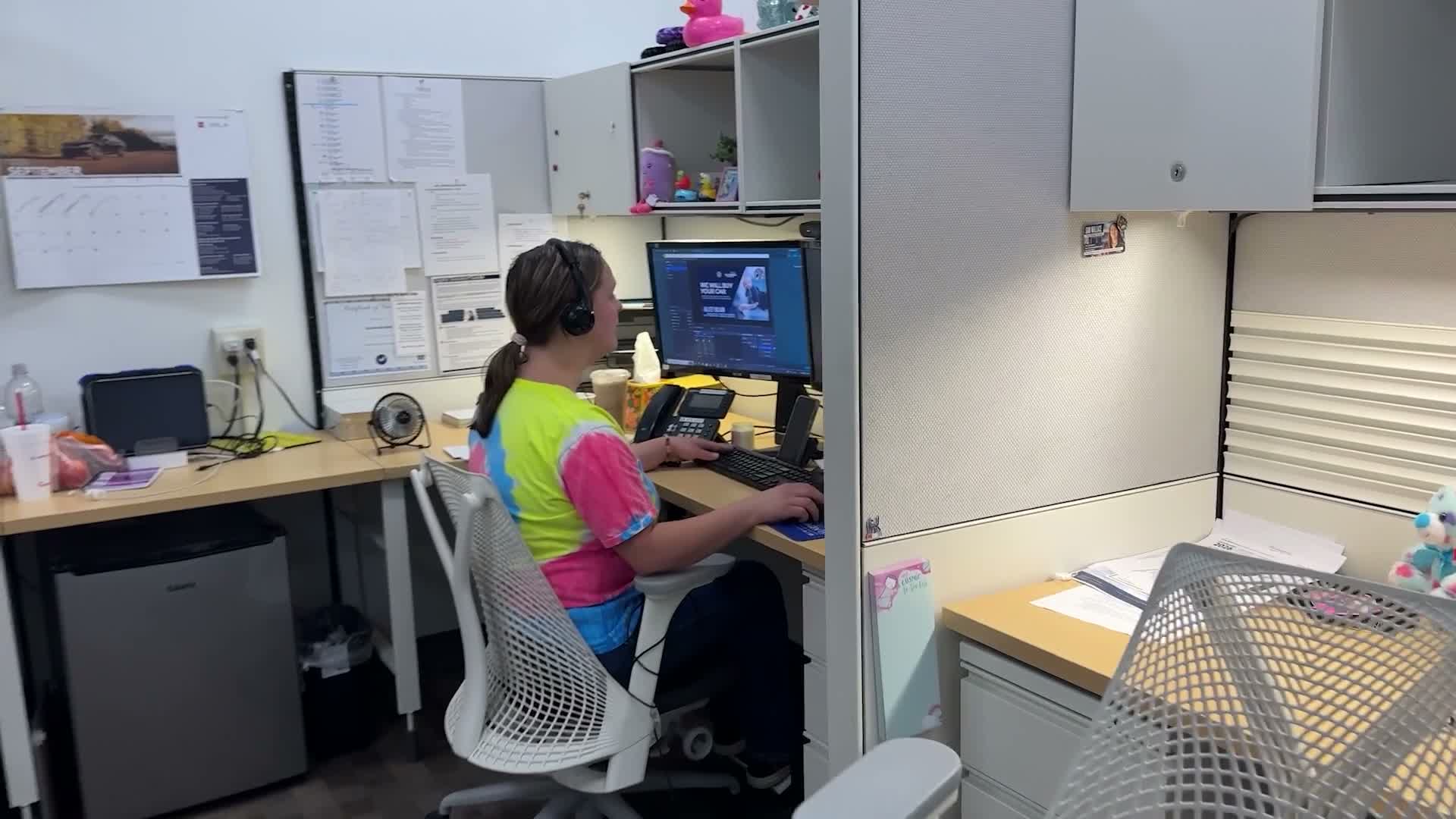 Video frame: an agent in a headset works at a cubicle desk, facing her monitor, on the BDC floor.